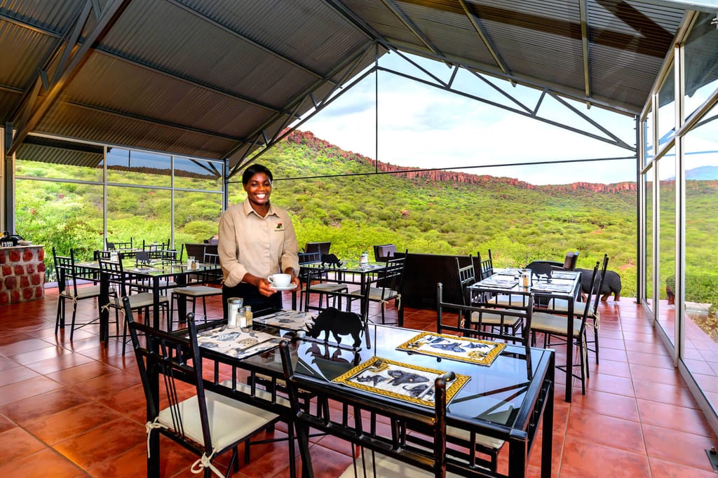 Überdachtes Restaurant mit gedeckten Tischen und Blick auf grüne Hügel und roten Felsgrat