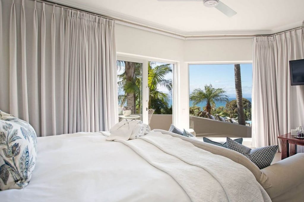 Helles Schlafzimmer mit Bett und Meerblick durch offene Fenster, Palmen auf der Terrasse sichtbar.