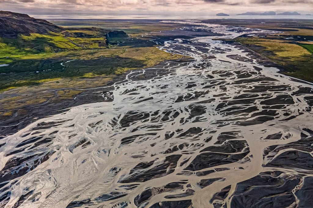 Luftbild eines verzweigten Schotterflusses mit hellen Wasserläufen und dunklen Sedimentflächen unter bedecktem Himmel.