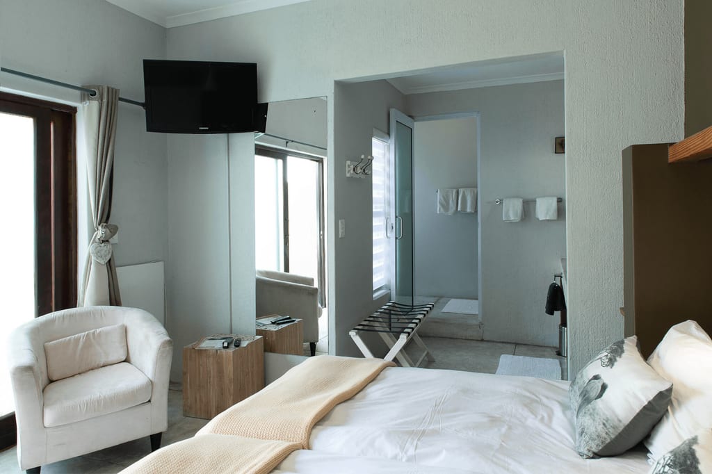 Hell eingerichtetes Hotelzimmer mit Doppelbett, Sessel und offenem Bad mit Glasdusche.