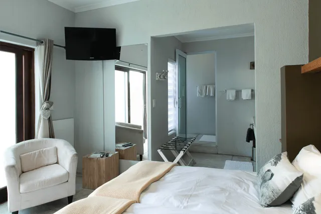 Hell eingerichtetes Hotelzimmer mit Doppelbett, Sessel und offenem Bad mit Glasdusche.