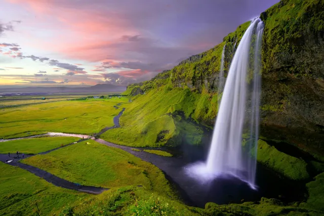 Hoher Wasserfall an moosbedeckter Felsklippe über grüner Ebene bei Abenddämmerung in Island.