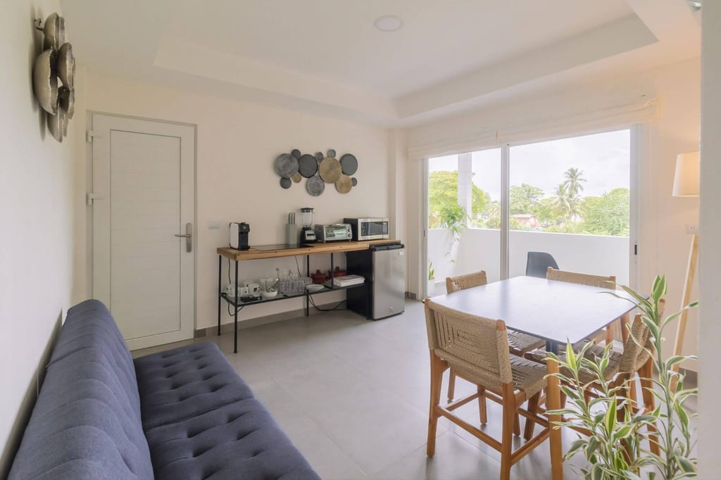 Helles Wohn- und Esszimmer mit Esstisch, Sofa und Sideboard vor Balkon mit Grün und Palmen.