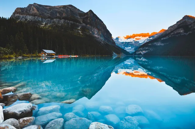 Türkisfarbener Bergsee mit gespiegelteten Bergen, Bootshütte und glatten Ufersteinen im Vordergrund.