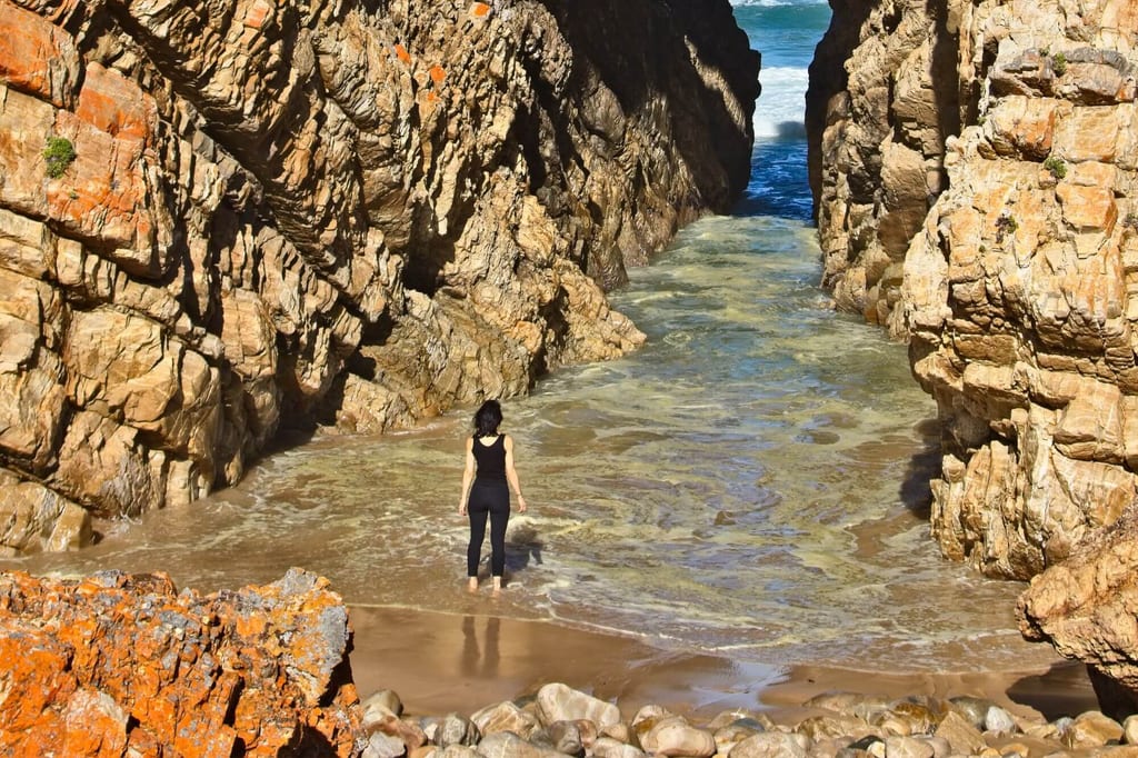 Person steht barfuß am sandigen Strand zwischen hohen, orange-braunen Felsklippen und schaut auf einen schmalen Meerkanal.