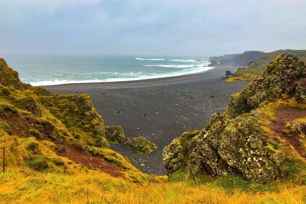 Blick von einer grasbewachsenen Klippe auf schwarzen Sandstrand und welliges Meer unter bewölktem Himmel.