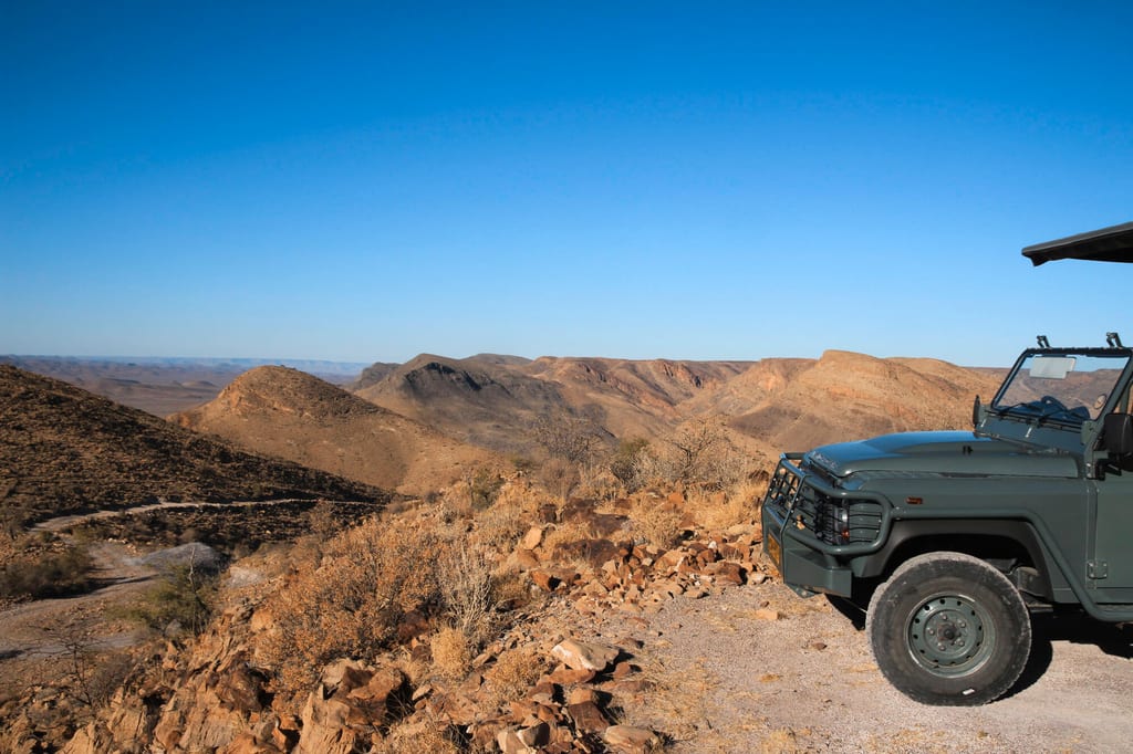 Safari-Fahrzeug am Rand einer trockenen, felsigen Berglandschaft unter klarem blauem Himmel.