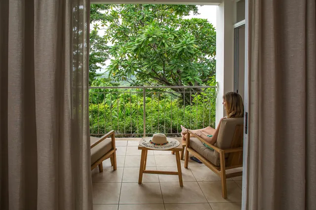 Frau sitzt entspannt auf Balkonstuhl und blickt auf dichten grünen Garten, Blick durch geöffnete Vorhänge.