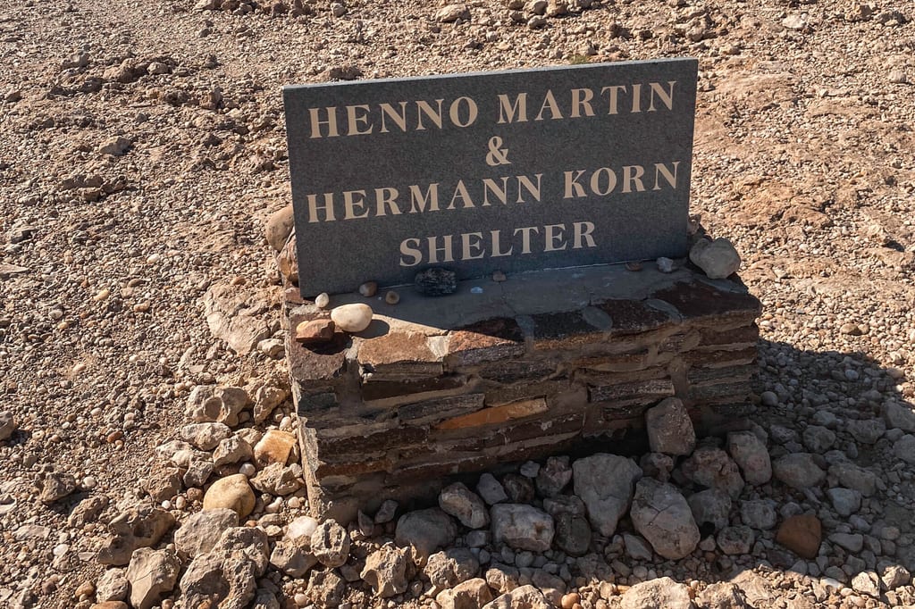 Gedenktafel mit der Aufschrift HENNO MARTIN & HERMANN KORN SHELTER auf gemauertem Sockel im sonnigen Schottergelände.