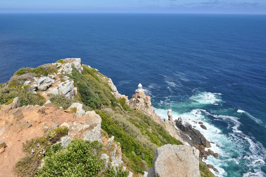 Blick von einer felsigen Steilküste auf einen kleinen Leuchtturm und das offene Meer bei sonnigem Wetter.