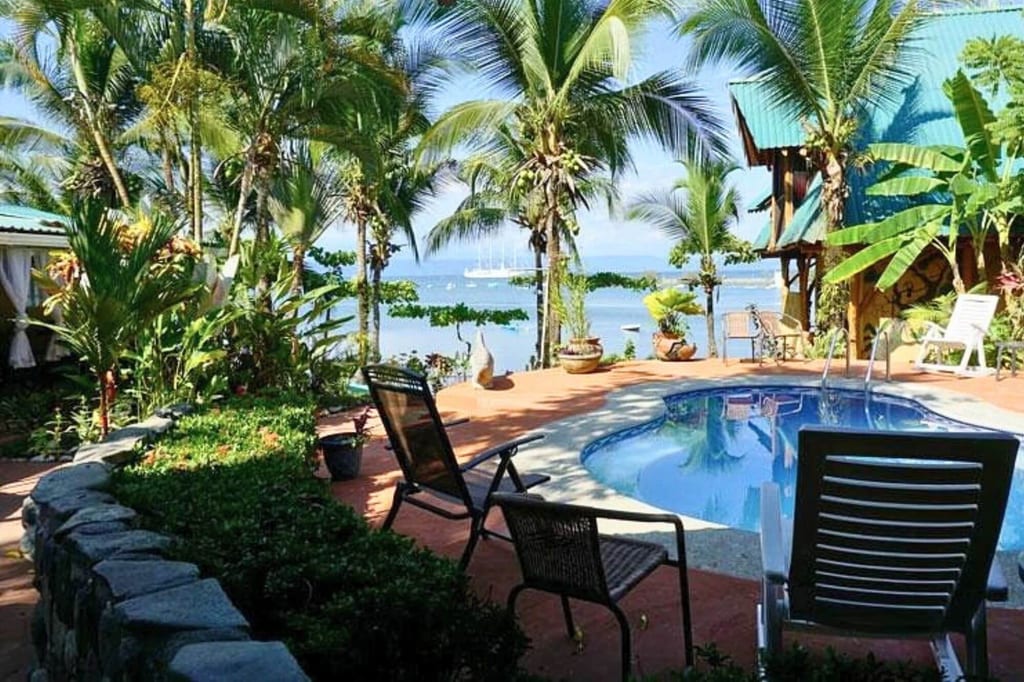 Blick von einer Poolterrasse mit Liegestühlen durch Palmen auf das Meer und Segelboote.