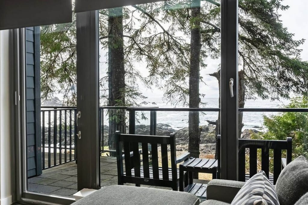 Innenraumblick durch Glas auf Terrasse mit zwei Holzstühlen und felsige Küste im Hintergrund, bewölkter Tag.