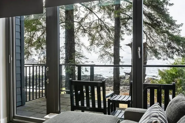 Innenraumblick durch Glas auf Terrasse mit zwei Holzstühlen und felsige Küste im Hintergrund, bewölkter Tag.