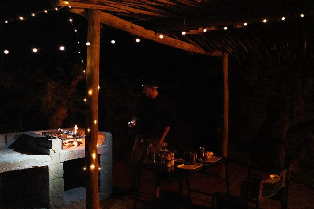 Nächtliche Außenküche mit Lichterketten, gemauerter Grill und einer Person am Tisch.