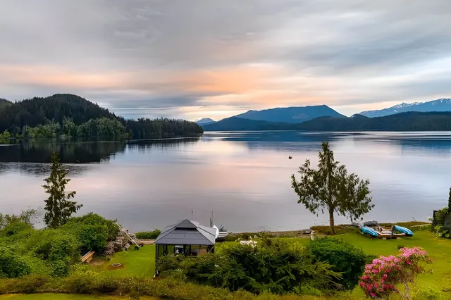 Dämmerungsblick über ruhigen See mit Pavillon, Booten und Bergen im Hintergrund, pastellfarbener Himmel