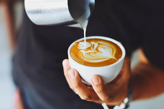Nahaufnahme: Hand gießt Milch in Tasse mit Latte-Art.