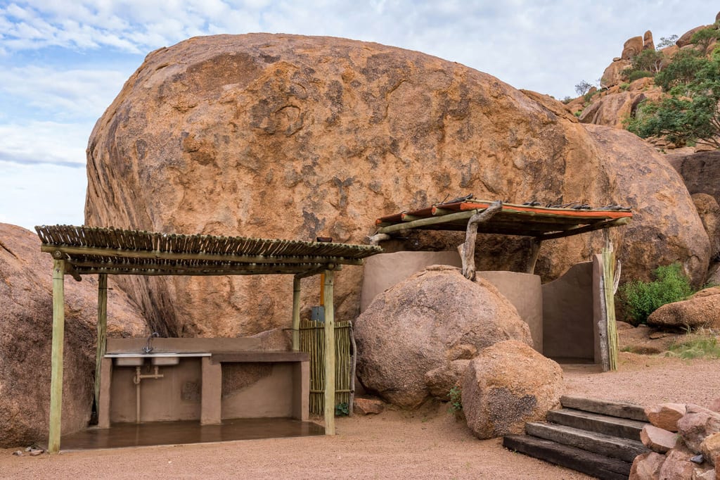 Außenanlage mit Waschbecken vor großen runden Granitfelsen und Holzüberdachungen bei bewölktem Himmel.
