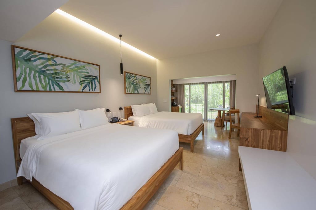 Helles Hotelzimmer mit zwei Doppelbetten, Holzmöbeln und großem Fenster zum Gartenblick.