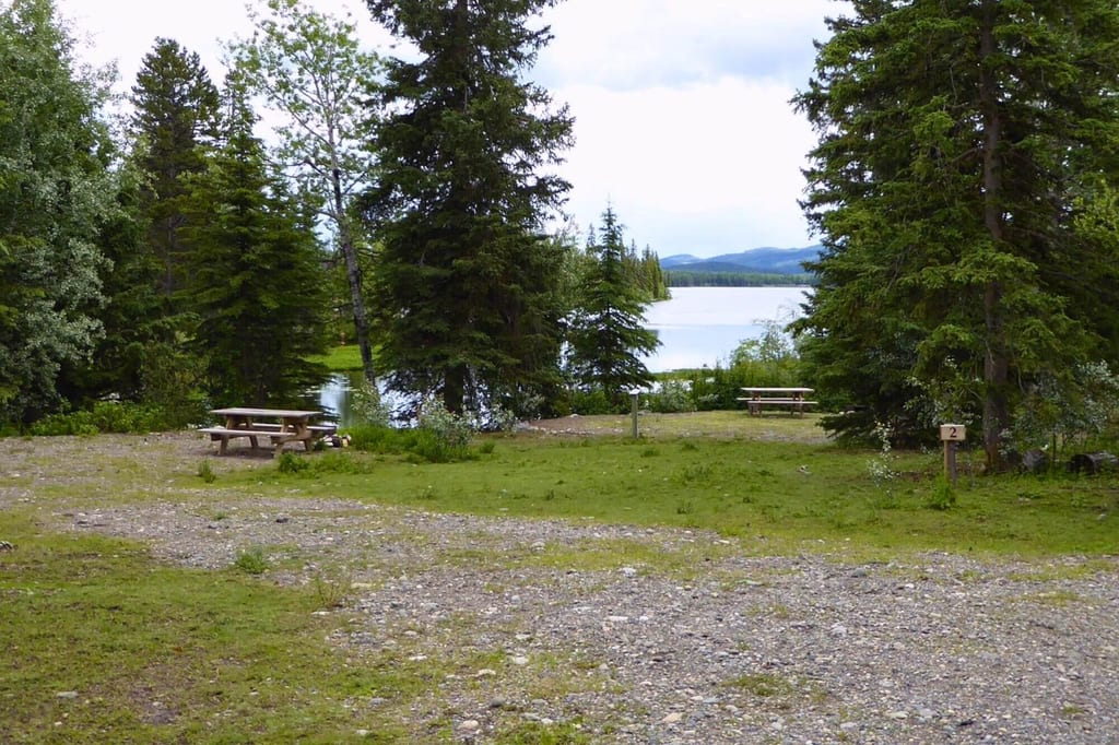 Schotterplatz mit zwei Picknickbänken, dahinter See und bewaldete Hügel bei bewölktem Himmel.