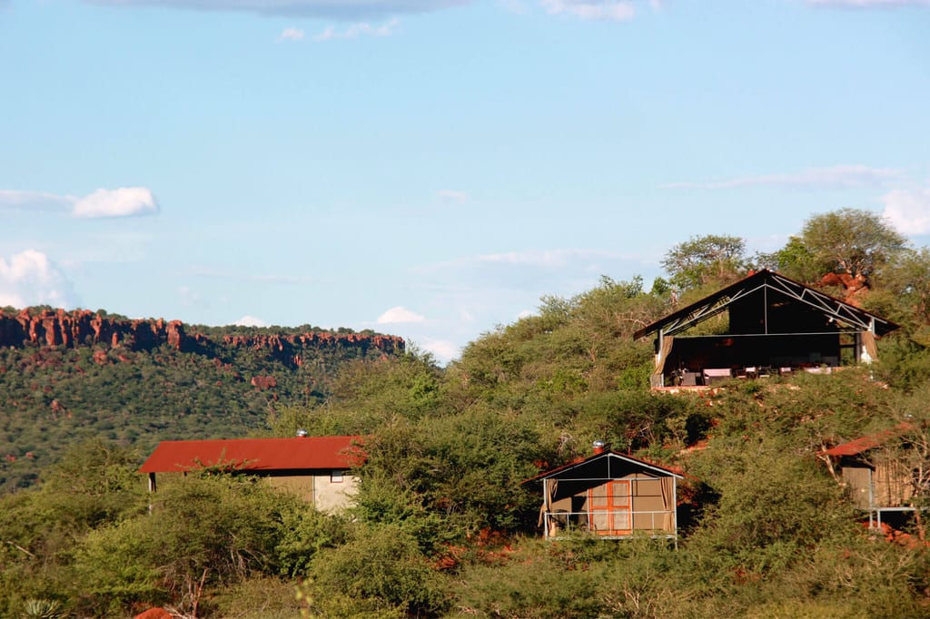 Safari-Lodges auf buschigem Hügel mit roten Felsklippen im Hintergrund, klarer Nachmittagshimmel.