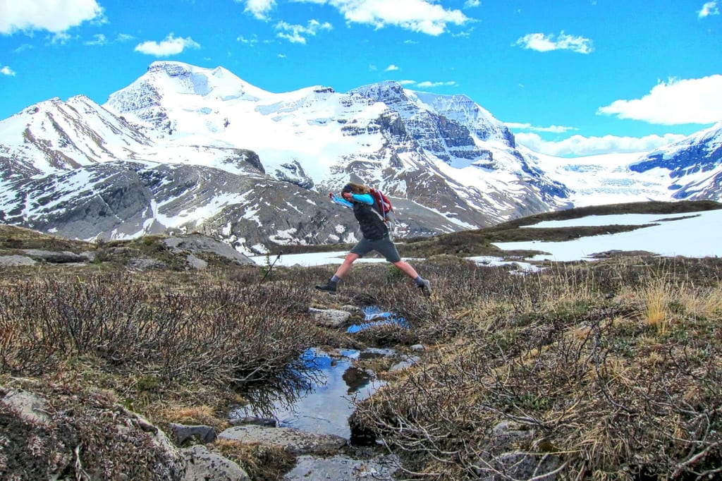 Wanderer mit Rucksack springt über Bach in alpiner Berglandschaft mit schneebedeckten Gipfeln.
