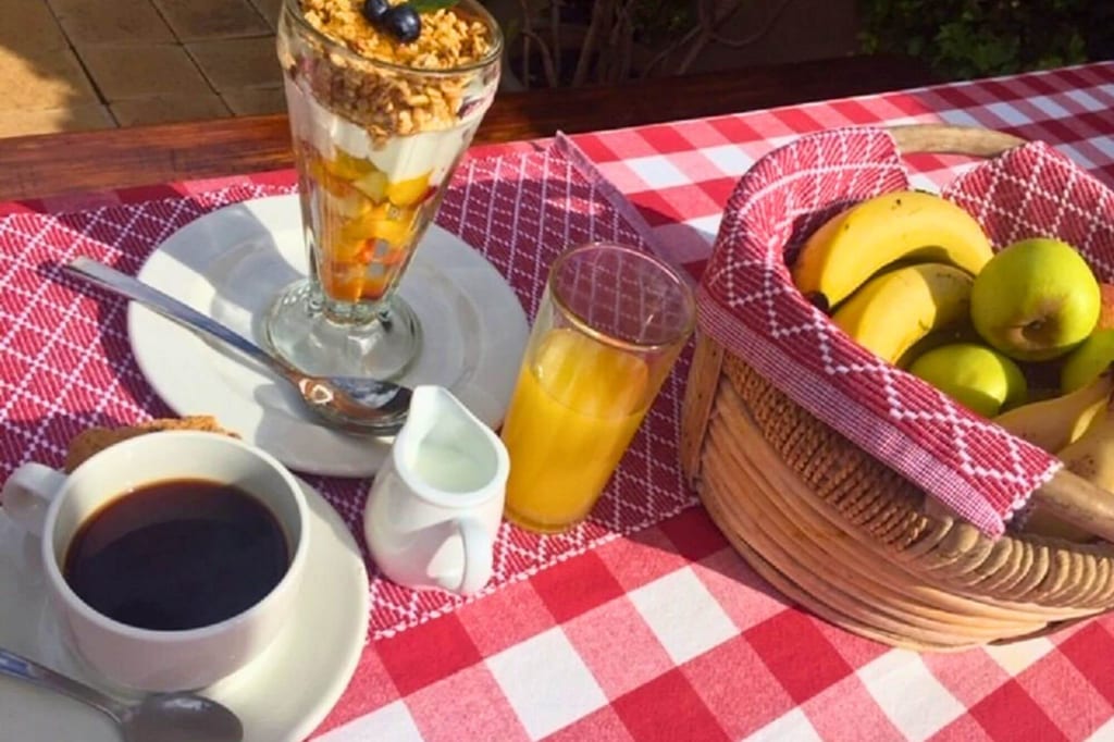 Frühstückstisch mit Kaffee, Joghurtdessert, Orangensaft und Obstkorb auf rot-weißer Tischdecke.