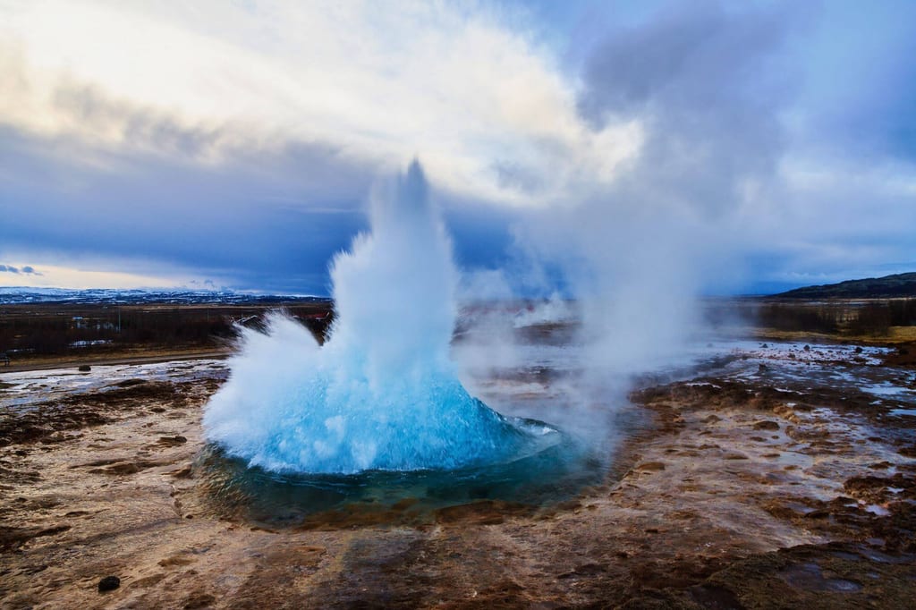 Geysir bricht aus, blaue Wassersäule und aufsteigender Dampf über kargem Boden