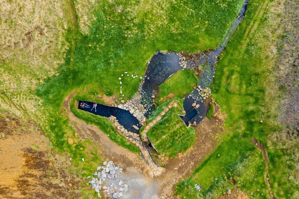 Vogelperspektive auf grasige Landschaft mit Bach, Felsbecken und zwei Personen im Wasser.
