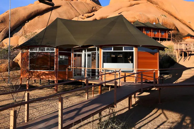 Holzsteg führt zu einer orangefarbenen Lodge vor großen Granitfelsen im warmen Nachmittagslicht.