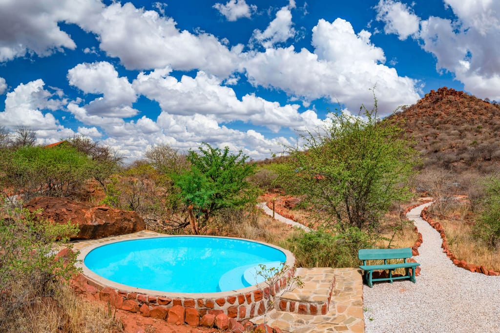 Rundes türkisfarbenes Pool mit Steinrand in trockener Buschlandschaft unter blauem Himmel und weißen Wolken