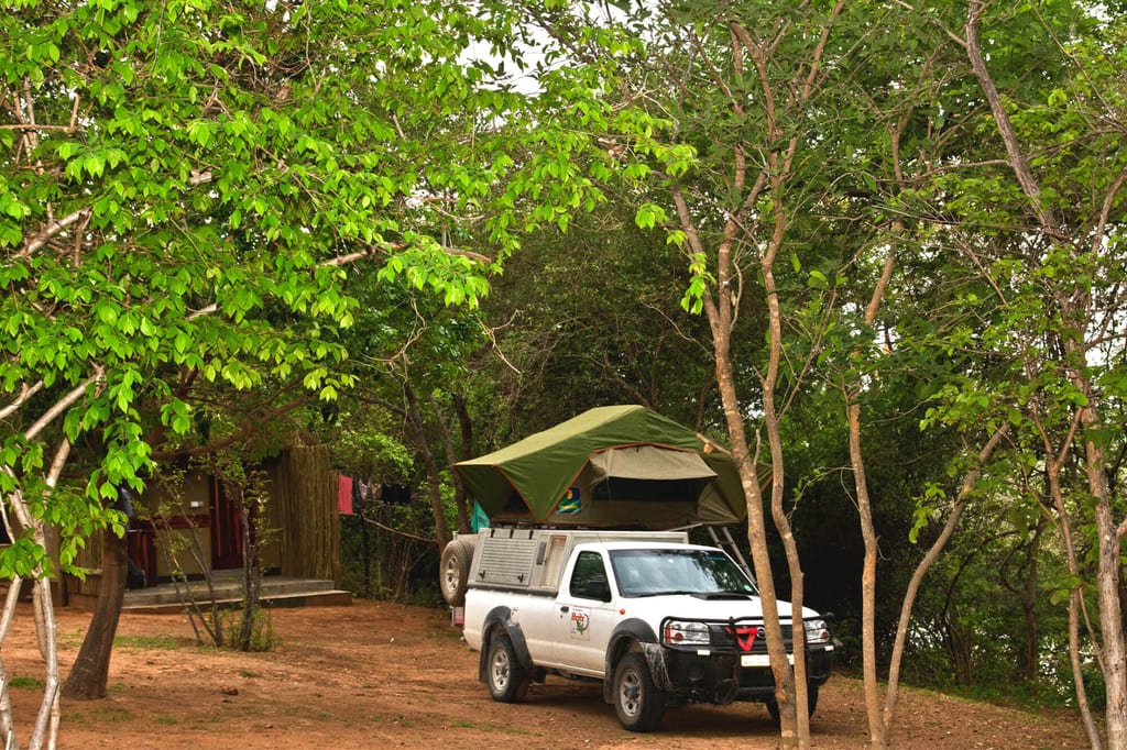 Weißer Pickup mit aufklappbarem Dachzelt auf schattigem Waldcampingplatz neben Holzhütte